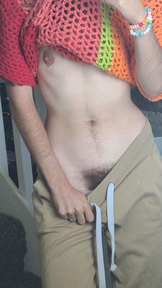 Adriiann stands wearing a red, pink, green, and orange stripped crochet net top, pulled up to reveal a nipple, and tan sweat pants that are being pulled down to reveal a lush bush. There's a slight tan on their torso, and their arm is noticeably more tan than their stomach