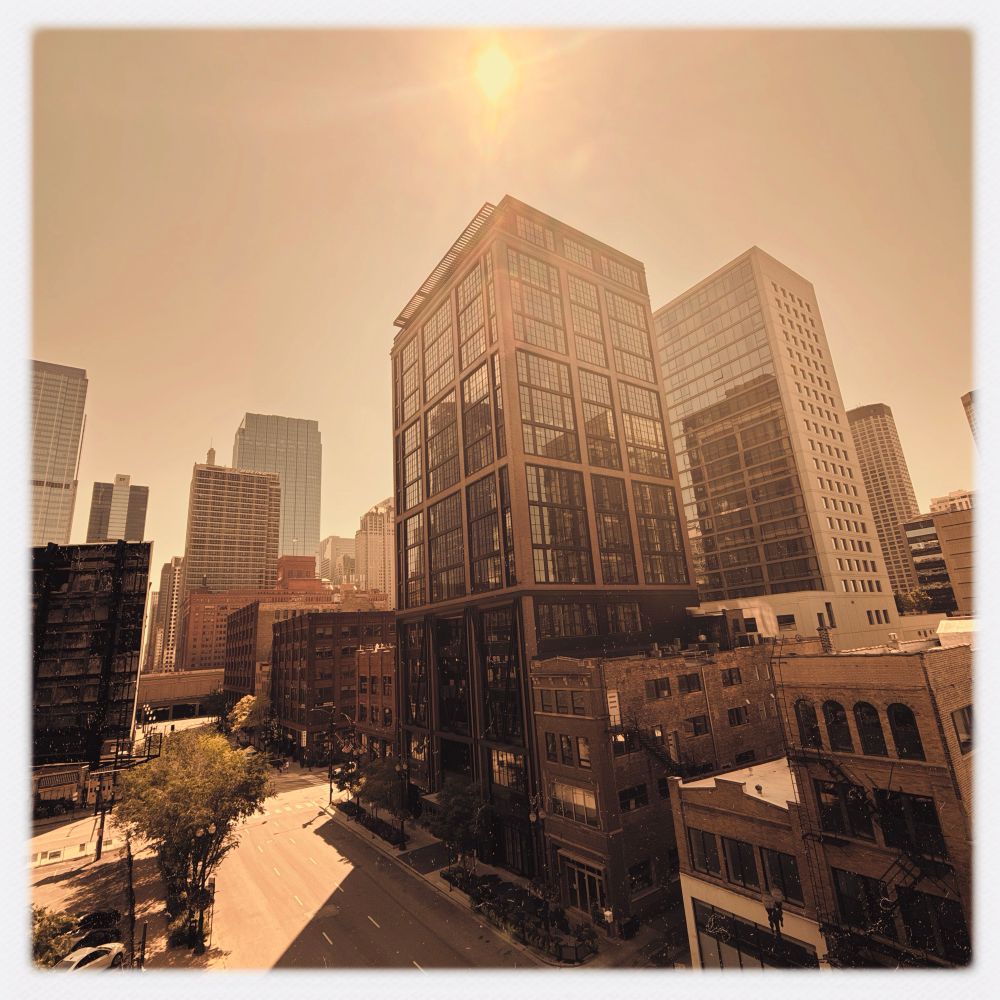 A cityscape featuring modern skyscrapers and historic buildings under a warm, sunlit sky. The street is partially visible, with trees lining the sidewalk. The image has a warm, vintage tone.