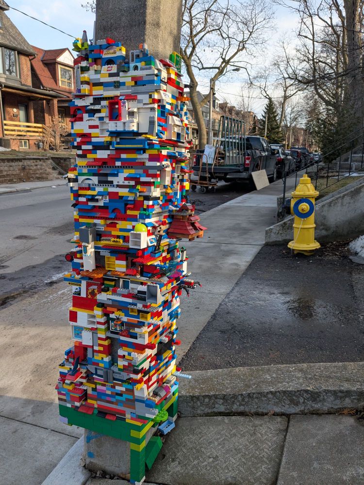 A utility pole on a residential street encased in an intricate, colourful LEGO sculpture about 3 or 4 feet high 