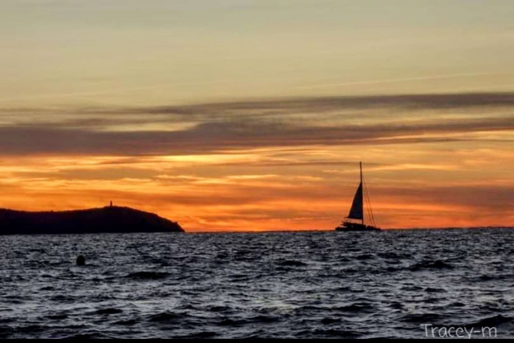 Silhouette of a sailboat gently gliding on water during a vibrant sunset with orange and purple hues, overlooking a distant island.