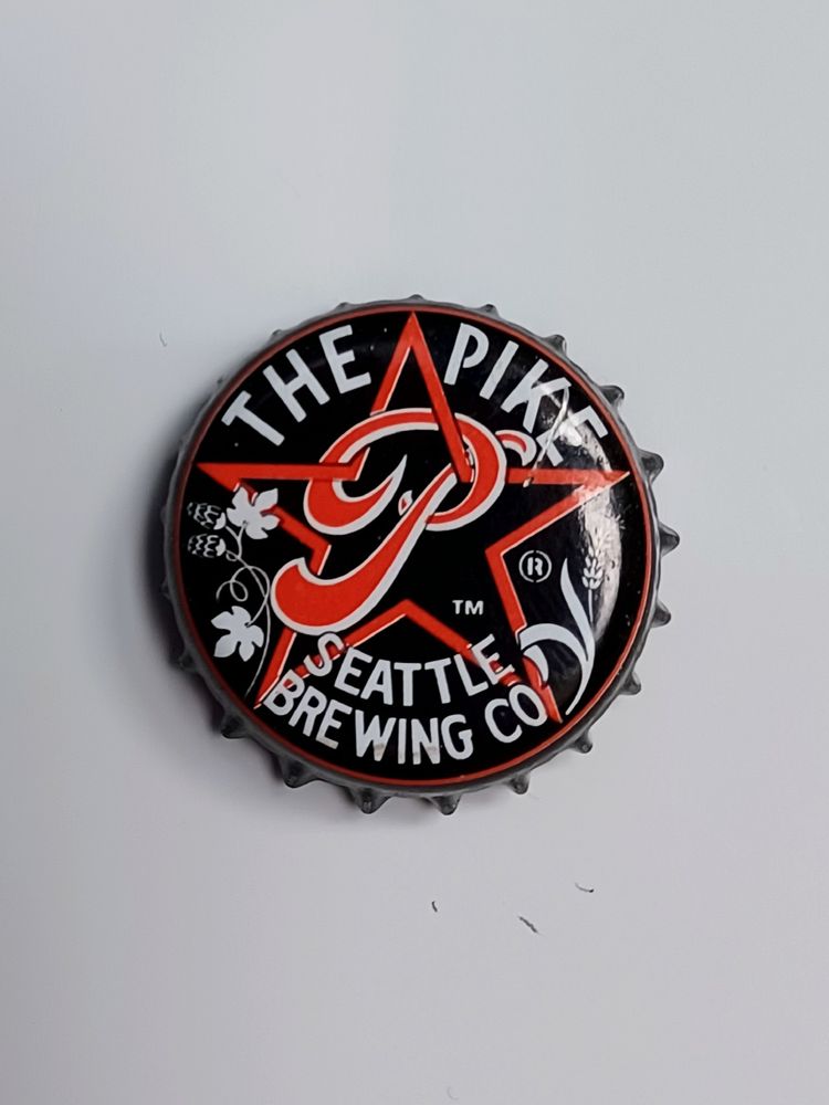 Crown cap against white background. Cap is black with white text and red logo. Logo is a star with a stylized P, with hops and barley plants adorning it. Text reads "The Pike Brewing Co, Seattle."