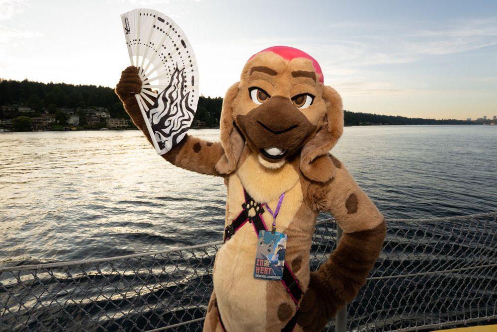 Killa' DeVille (A fursuit of a Rabbit with a brown fur pattern of a hyena and a hot pink bandana.) standing on a boat in a black and pink body harness and holding a white fan with a flame design. 