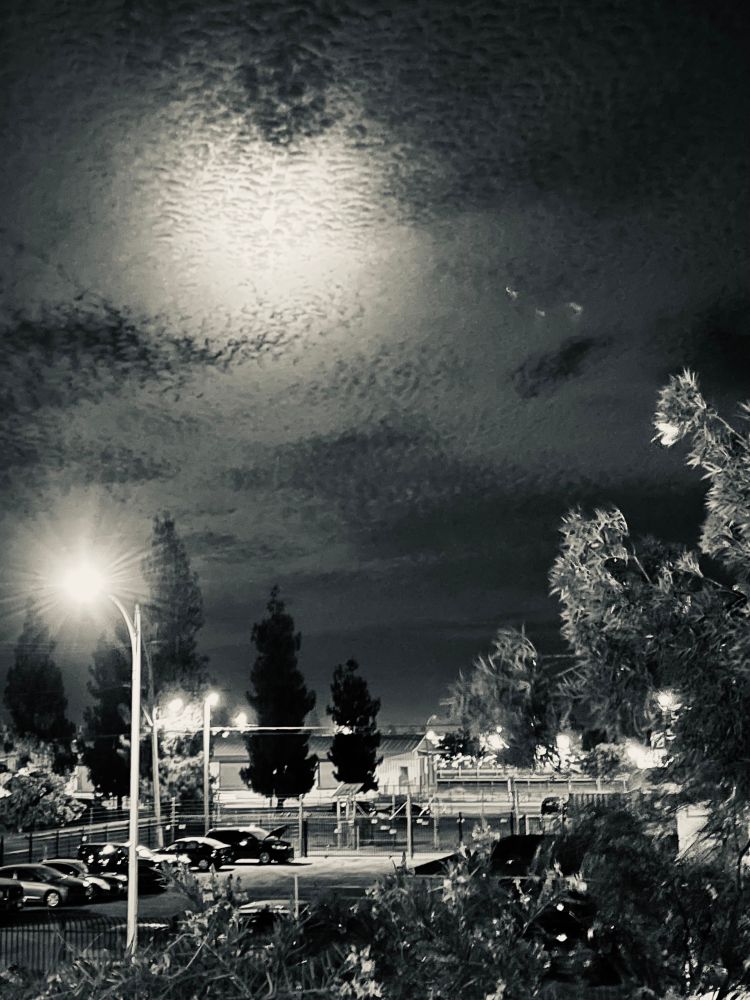 Photo shows small full moon light behind undulating rivulets of clouds with urban street lights and a parking lot lined by tall trees. 