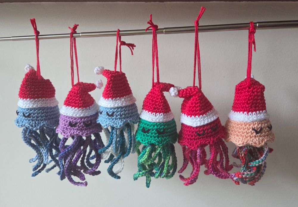 Six crocheted jellyfish with red Santa hats and multicoloured tentacles, hanging from a metal rod. Shown against a white background. Colours from left to right: ice blue with multicoloured blue tentacles; lilac; ice blue with cream, ice blue and teal tentacles; green; red; and salmon