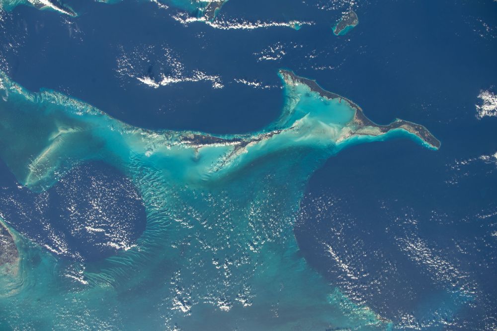 The image shows irregular shaped long islands with a torqouise coloured sea next to them. Some parts of the ocean are deep blue. There are also some small clouds scattered over the area. 