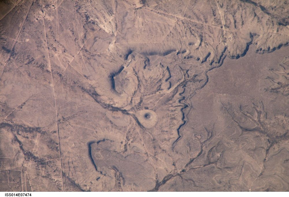 The image shows a brownish landscape with peculiar shapes and a kind of eye in the centre of the image. The peculiar shapes are the borders of flat soft sediments capped with a layer of harder rock which resists erosion. The soft layers can get eroded sideways and this causes flat plains and isolated hills with steep borders. Images like these often provide an optical illusion. The dark lines in the image are the steep borders which are in shadow. The sunlight comes from the right and the mesas are elevated over the surrounding terrain. Often however the brain sees a reverse relief and lets the mesa look like pits or depressions in stead of mounds and elevated ground. 