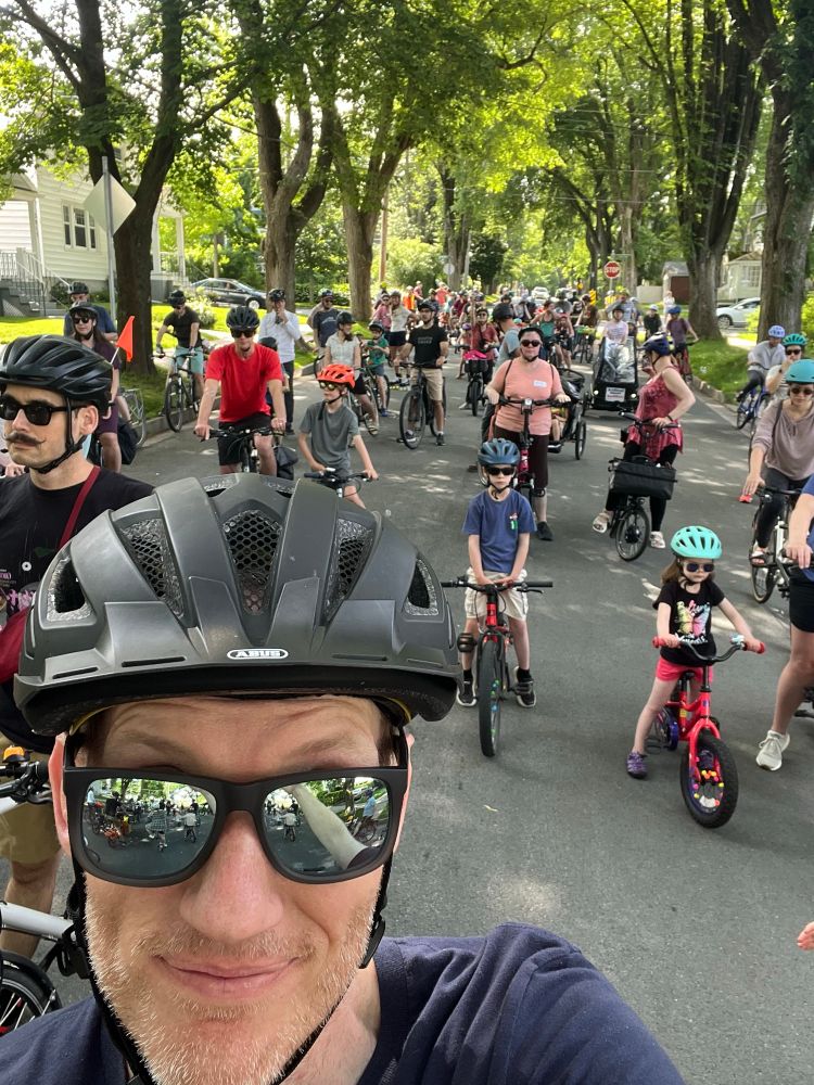 A selfie of me, a white man wearing sunglasses and a bike helmet. Behind me are many more people on bikes on a street in Halifax 