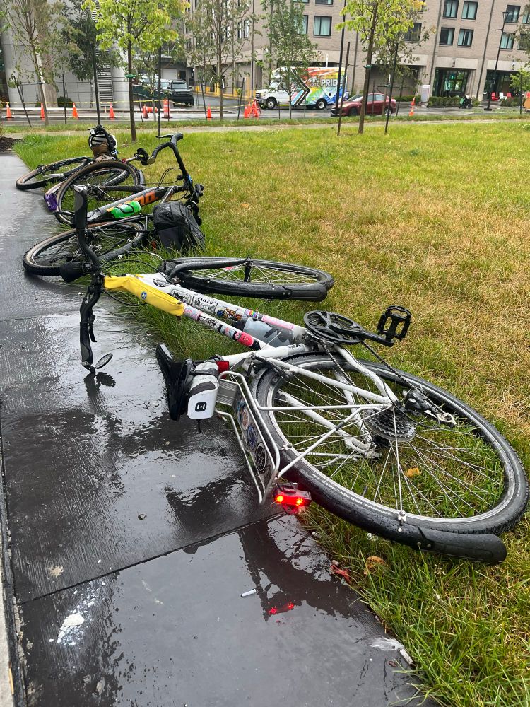 Bikes laid on the grass, the concrete nearby is wet from a bit of rain 