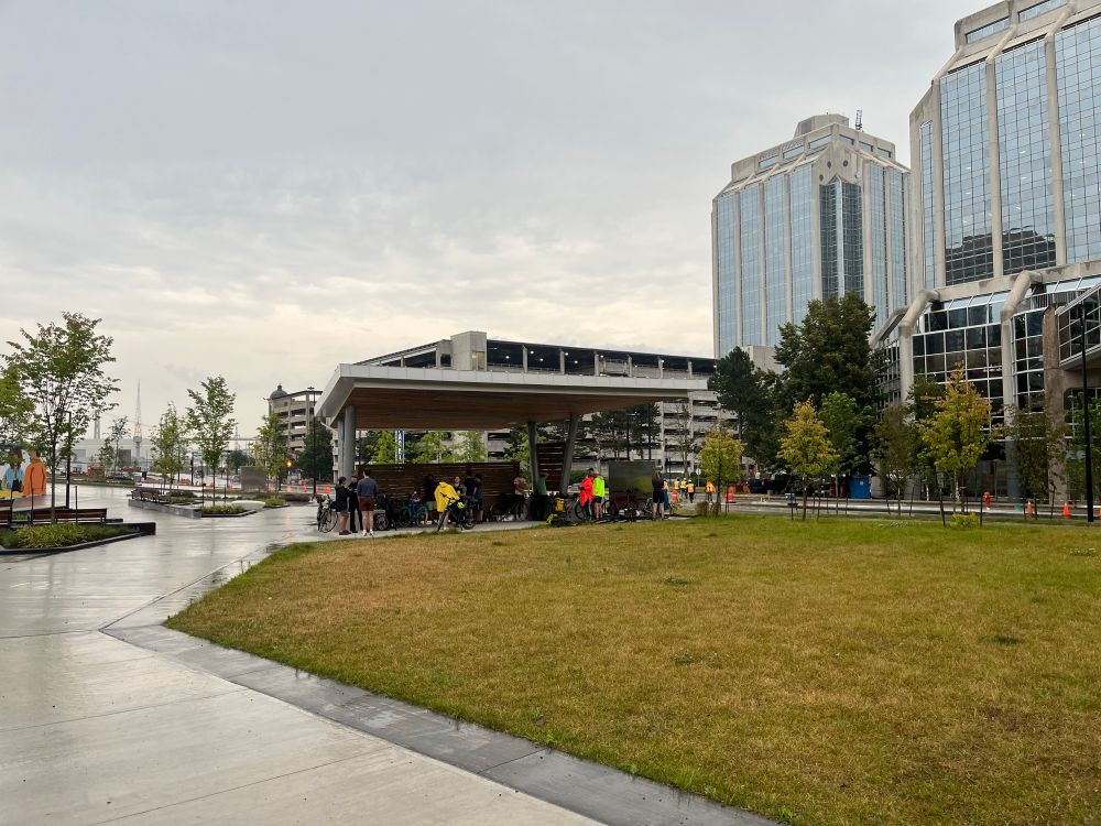 A view of the performance stage as part of the new Cogswell area in Halifax. People and bikes are around and under the overhang. Purdy’s Wharf buildings are visible to the left.