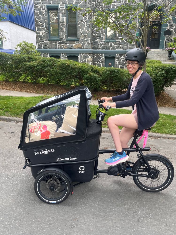A white woman on a trike with a bucket loaded with supplies for the ride and a folding bicycle 