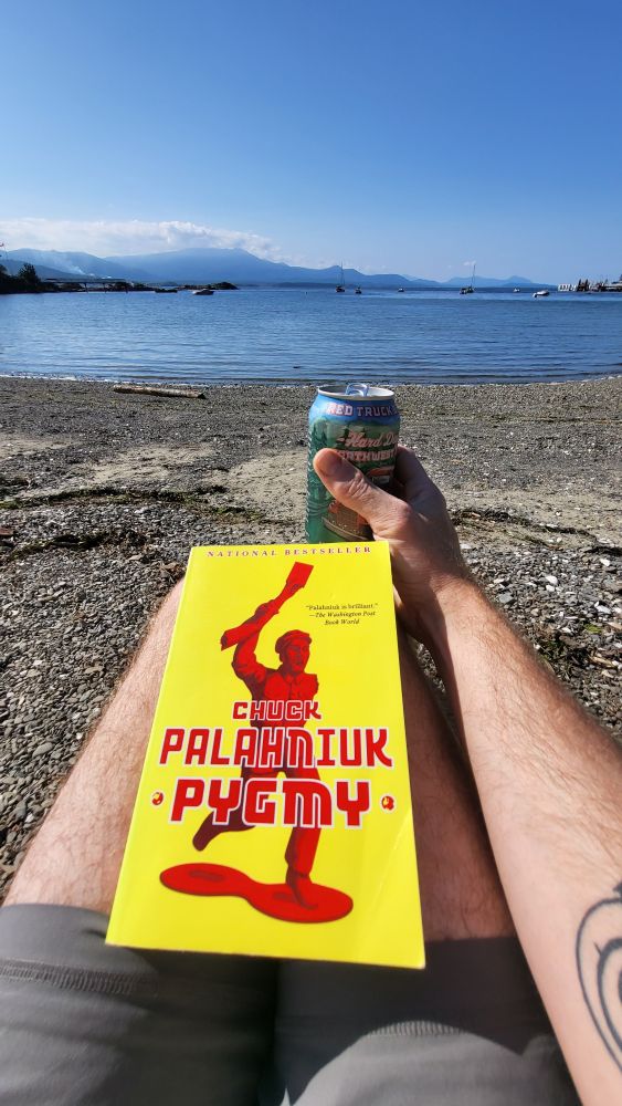 A copy of Pygmy by Chuck Palahniuk on my lap while I hold a beer overlooking the ocean on a pebble beach.