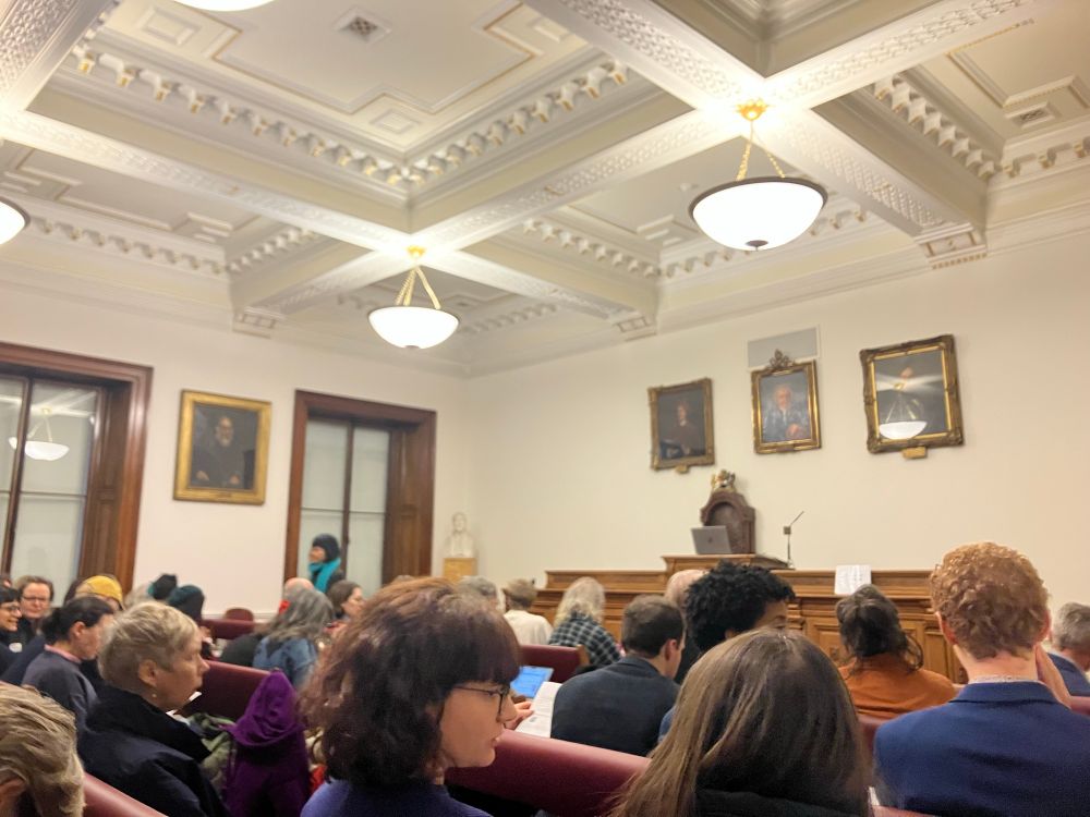 White walled room with patterned plaster ceiling, old portraits on the wall & a wooden platform at the front. The room is full of people. 