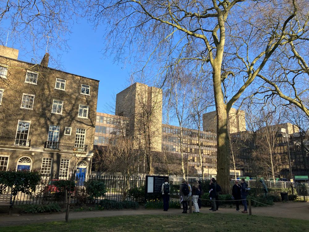 Landscape image. Park in the foreground. Large brutalist building behind. Set in front of a clear blue sky. 