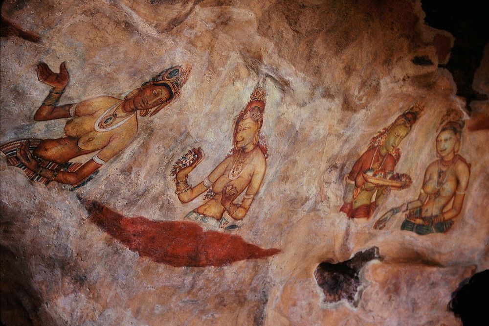 Frescos of voluptuous women painted on the Mirror Wall of Sigiriya. Four in shot. All wear elaborate crown-like headdresses and multiple necklaces. The first raises her right hand. The second holds a bunch of flowers in her right hand. The third holds a tray (containing flowers?), the fourth is gazing upon the tray held by the third, and herself holds things in her hands (probably also flowers). All four appear to be standing waist-deep in clouds.