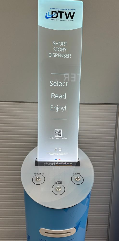 A tall stand in an airport corridor with signage to let patrons know that if they push a button, it will spit out a free short story on recycled paper for their enjoyment