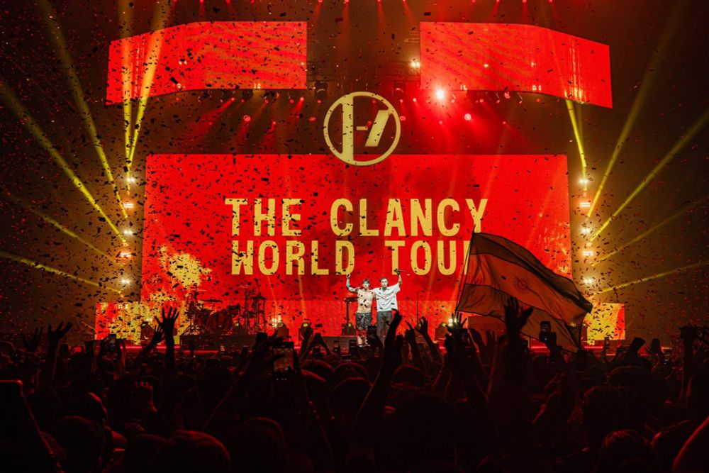 twenty one pilots onstage closing their first show in buenos aires, argentina. both stand side by side holding each other's shoulders, raising a hand to the public. a giant fire red screen in the back says "the clancy world tour" in yellow letters, their logo in yellow as well right above it. the people in the crowd have their hands raised, and an argentinian flag is visible at front row. 📸 dfallaccess