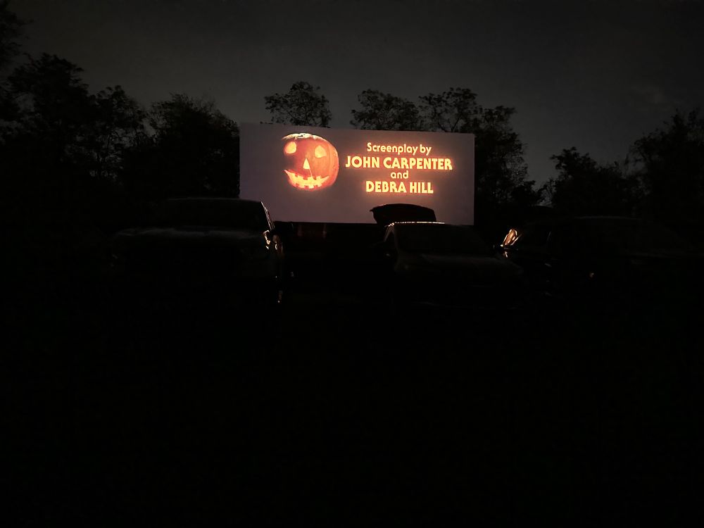 A night time photo of a drive-in screen displaying the opening credits of Halloween. The screen reads: Screenplay by John Carpenter and Debra Hill.