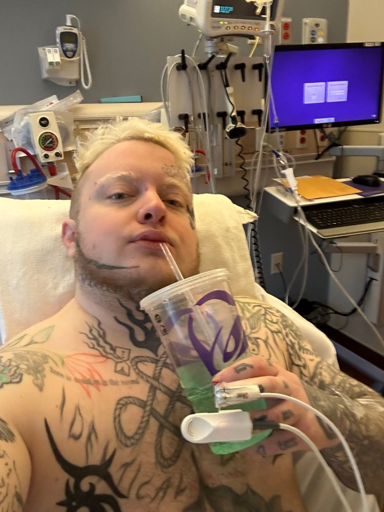 Eoin, a heavily tattooed white trans man, lays shirtless in a hospital bed after surgery. He still has monitors and IVs attached as he drinks a large Taco Bell Baja Blast. 