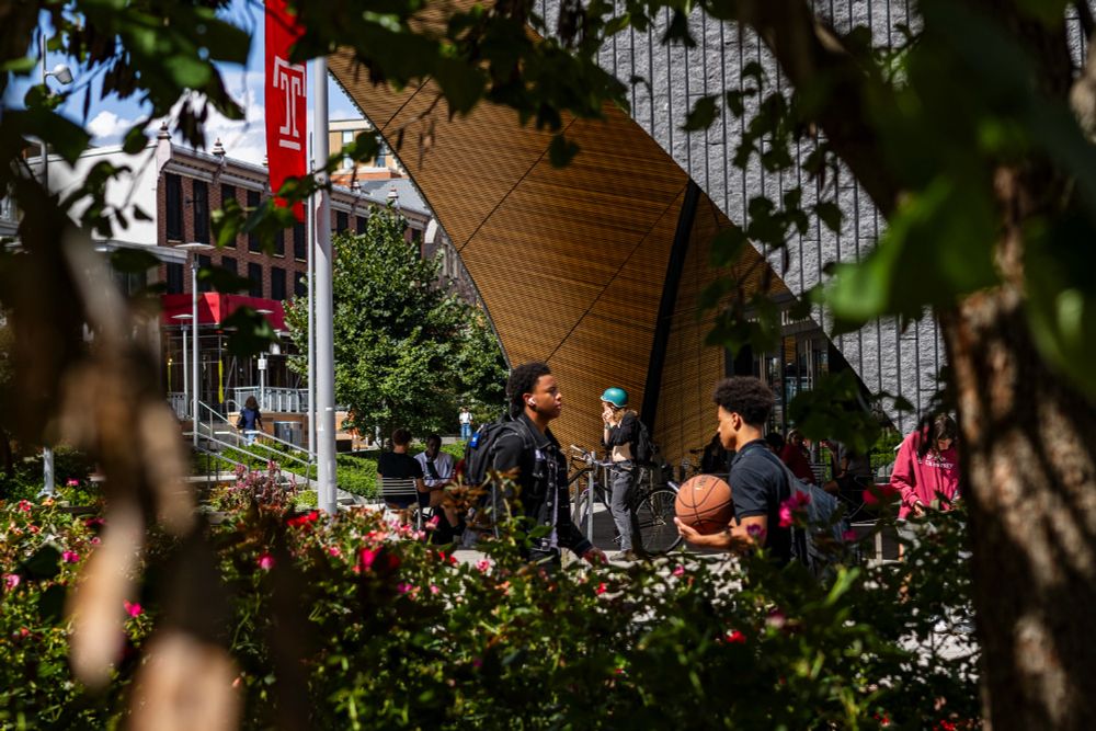 A group of students stand outside Charles Library with a large wooden arch and gray walls. One person is holding a basketball. A red flag with the white letter “T” hangs on a pole, and flowers and trees are in the front. Other people are sitting and walking nearby.