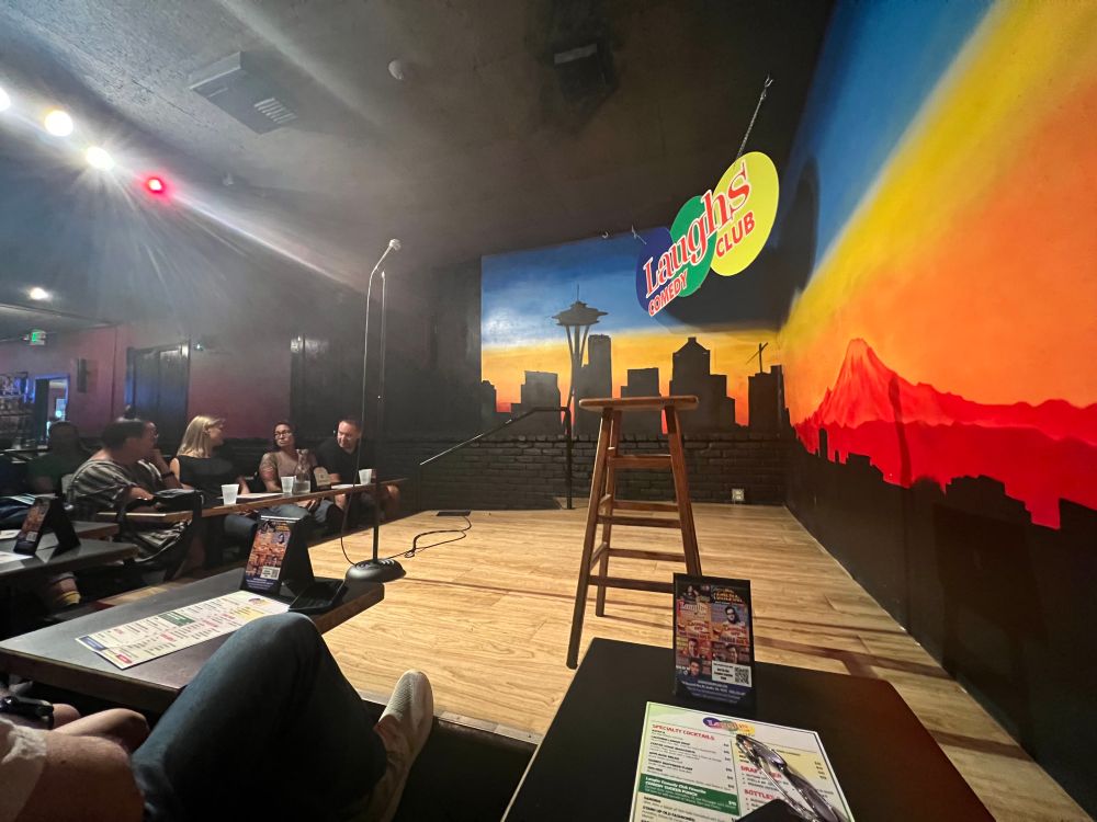 Comedy stage set with a stool and microphone. Sign reads “Laughs Comedy Club.” Mural shows Seattle skyline.