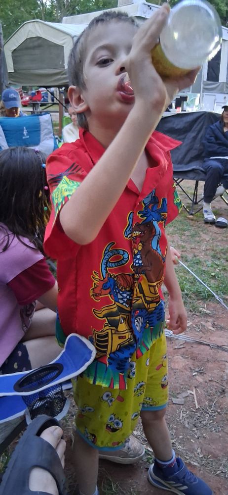 A young boy at a campsite in colourful clothing chugging the remnants of a bottle of raspberry cordial.