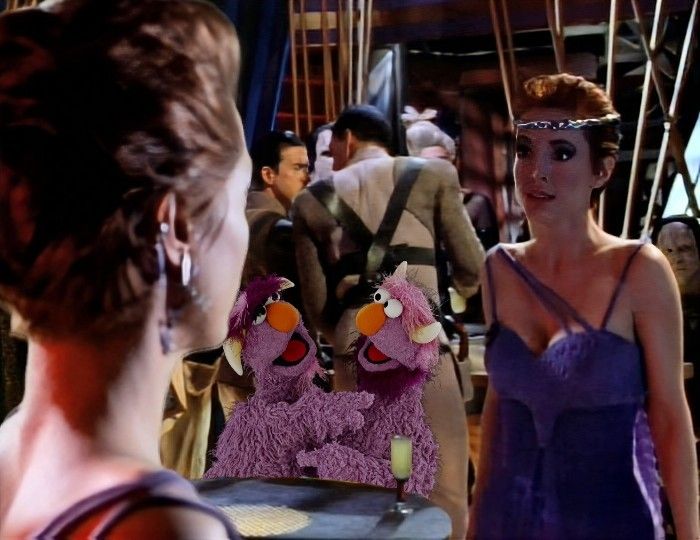In a scene from Deep Space Nine, Major Kira is aboard the mirror universe station run by her counterpart. In the foreground, on the right, we see the back of Prime Kira as she looks at Intendant Kira. The Intendant looks extremely miffed. Behind the Intendant, positioned between the two of them, sitting at a table is the purple Two-Headed monster from Sesame Street. 