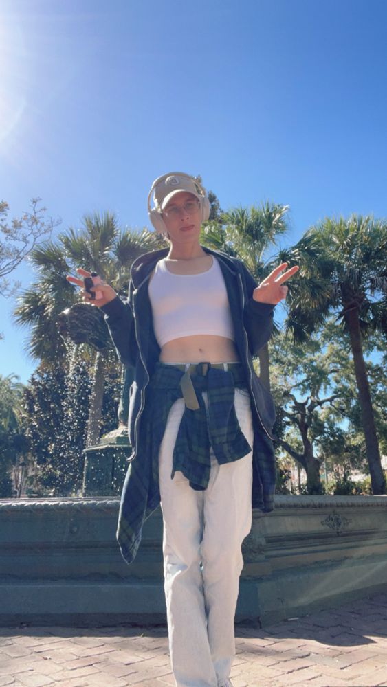 Brunette woman posing in front of a bronze fountain.Wearing blue jeans and a white tank crop with dad hat. 
