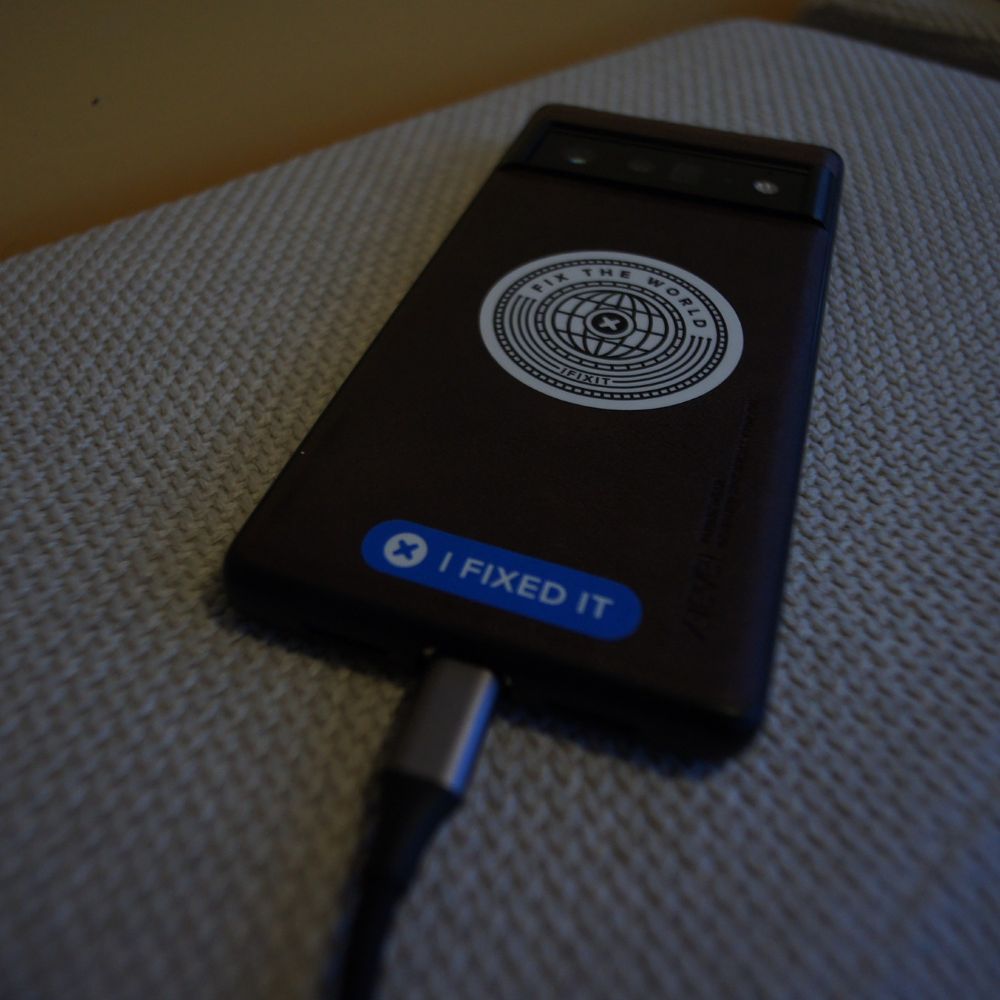Photo of a Google Pixel 6 Pro phone sitting face-down on a fabric couch. There are two stickers on the back: centred on the bottom is a rounded-rectangular blue sticker reading "I FIXED IT" with an Ifixit logo on the left. Near the centre of the phone is a circular sticker like a crest reading "FIX THE WORLD" and "IFIXIT" on the outside, framing a globe and an Ifixit logo.