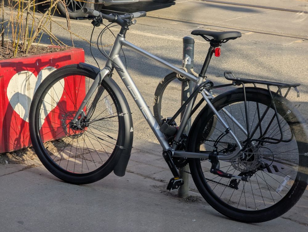 A shiny, silver hybrid bike locked up by the side of a street. In front of it is a concrete planter painted with a white-on-red heart; it holds spindly plants. The bike has splash guards, a pannier rack, and a VERY big lock attaching it to a bike post.