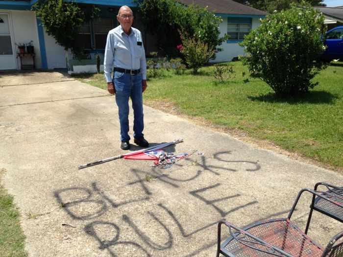 An old white man stands in front of a burnt Mississippi flag in his driveway. Nearby, the phrase "Blacks Rule" has been spray painted on the ground. The old man looks devastated at the damage to his driveway that he absolutely did himself.