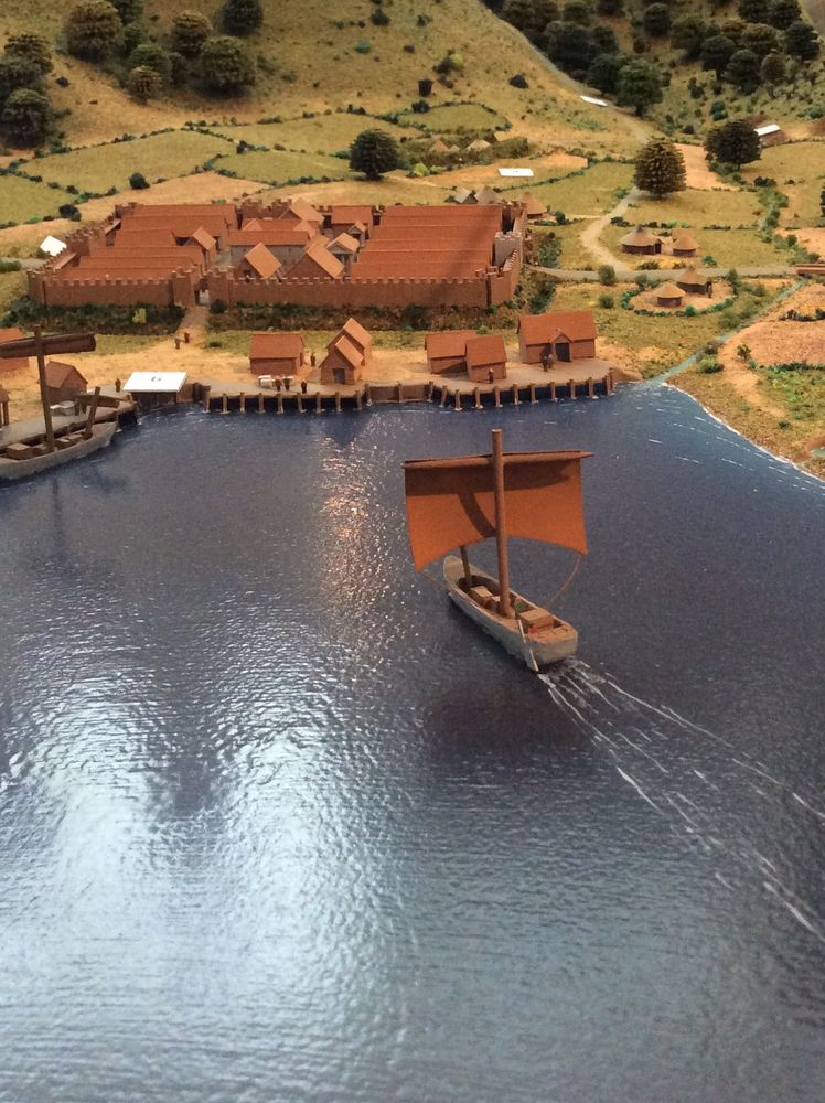 The diorama shows the sea in the foreground with a small boat with a red sail heading towards the harbour. There is a cluster of buildings around the harbour. Most of the buildings are within city walls. Behind these are the countryside. 