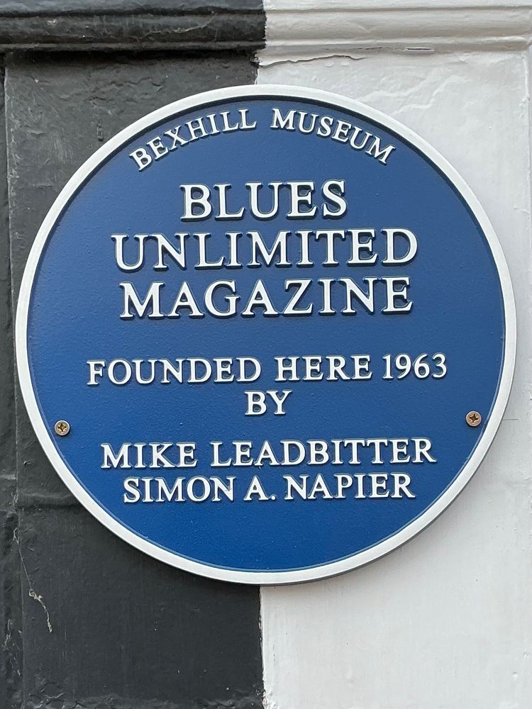The round blue plaque has a thin white border. The writing reads “Bexhill Museum. Blues Unlimited Magazine founded here 1963 by Mike Leadbitter (&) Simon A. Napier. “