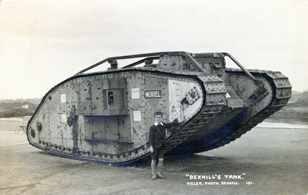 Black and white photograph of a tank as a postcard c.1920. "Bexhill's Tank." A boy stands next to a World War One tank on Bexhill's seafront.