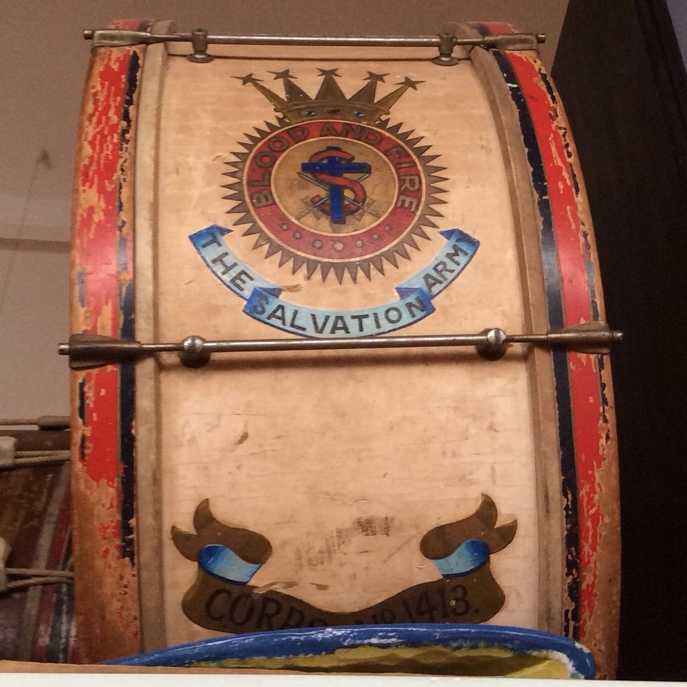 The side view of the drum. It is standing on its side. The text on the side of the drum reads “blood and fire. The Salvation Army.” There is a logo of an S wrapped around the cross. There are also 2 swords.  On top of the logo is a picture of a gold crown. 