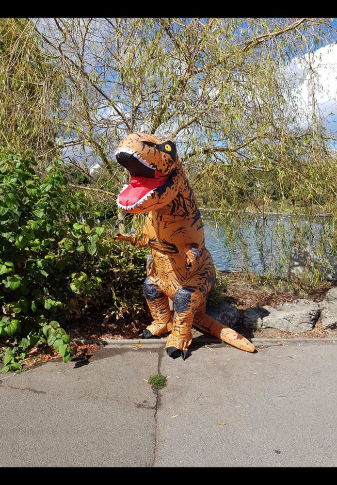 The photograph shows a person inside a various shades of brown dinosaur costume. He is standing in some foliage in front of the boating lake in Egerton Park, #Bexhill 