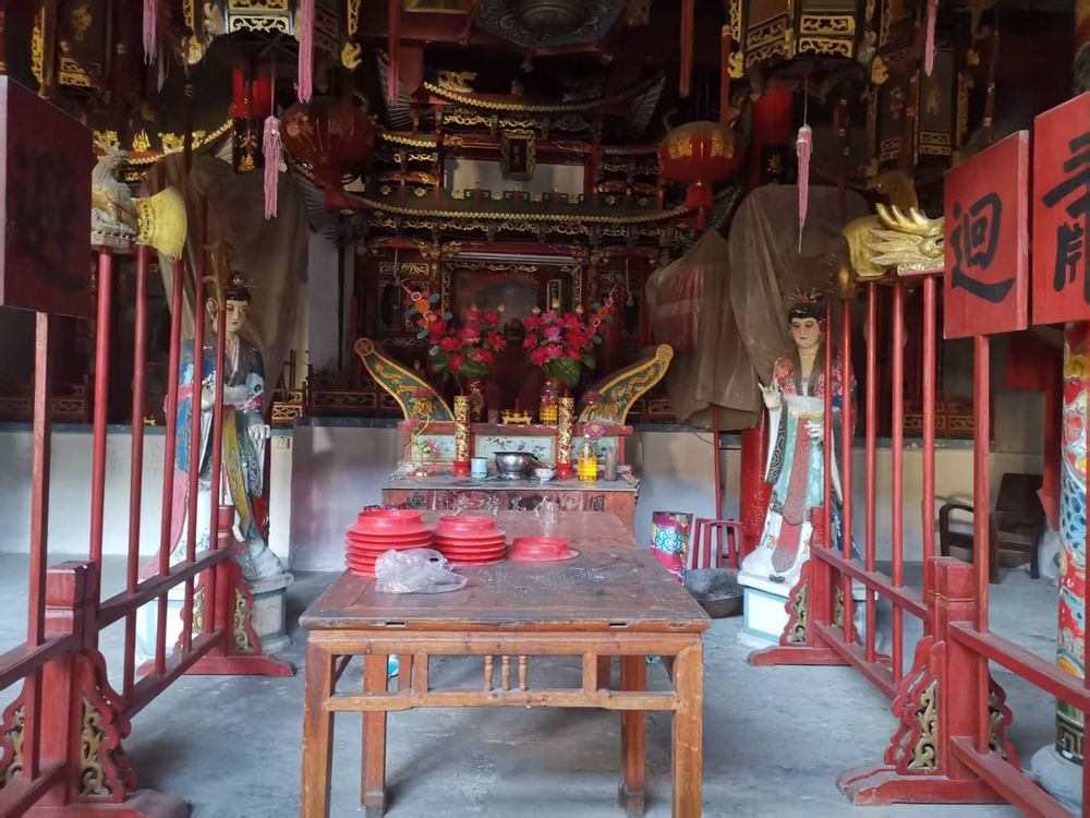 A central altar inside a Wenzhou Daoist temple, framed by vivid red railings, carved gold ornaments, and two elaborately painted guardian figures. Red offering bowls and temple decorations fill the wooden table in the foreground, showcasing local folk-art traditions.