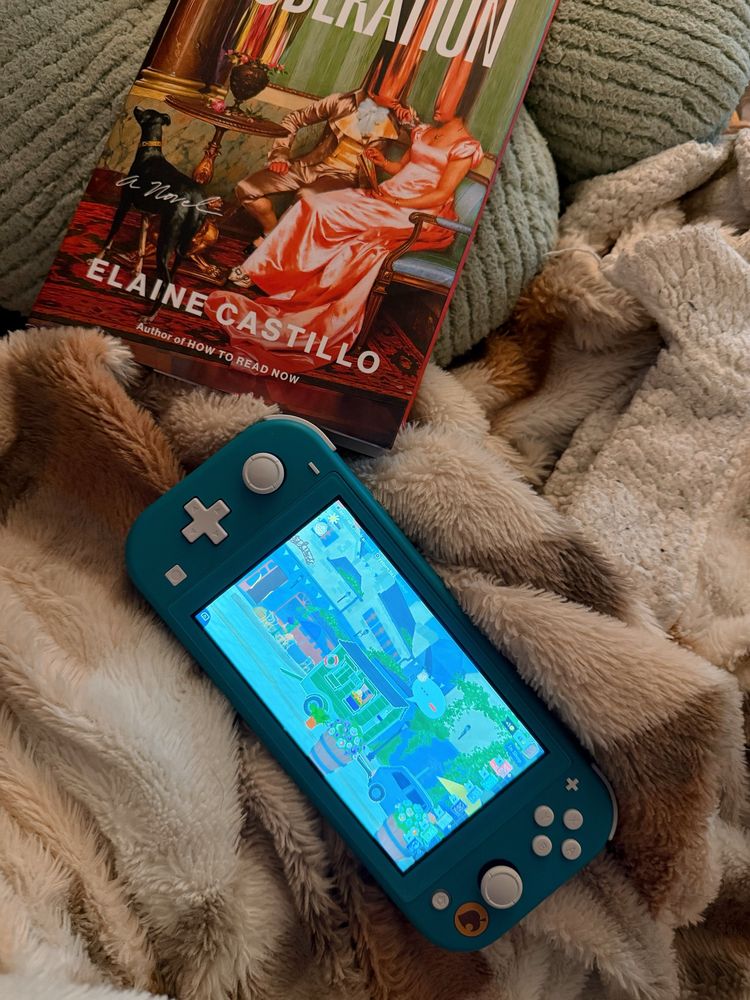 Picture of my current view, including the book Moderation by Elaine Castillo next to my turquoise Nintendo Switch, which currently displays a screen from the new game Tiny Bookshop