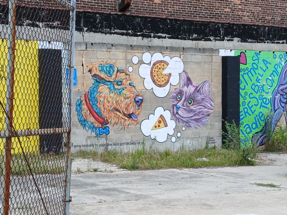 Mural on a concrete wall of a brown dog with a red collar thinking about a pizza with a slice removed and a gray cat thinking about a slice of pizza. The quote 
