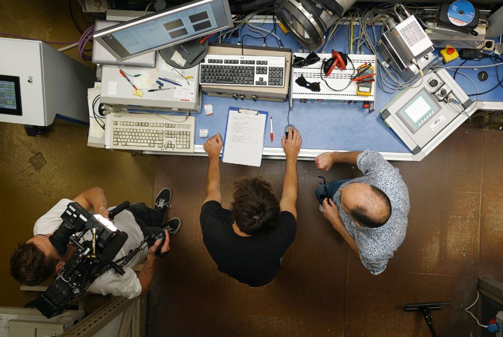 Bird's eye view of a desk with a computer, in front of which there are two men and a camera operator