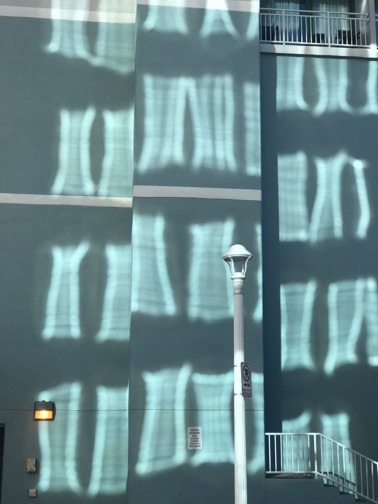 loose grid of reflected sunlit windows on a wall, with a central vertical lamp post and vertical railings at top and bottom right, taken this morning