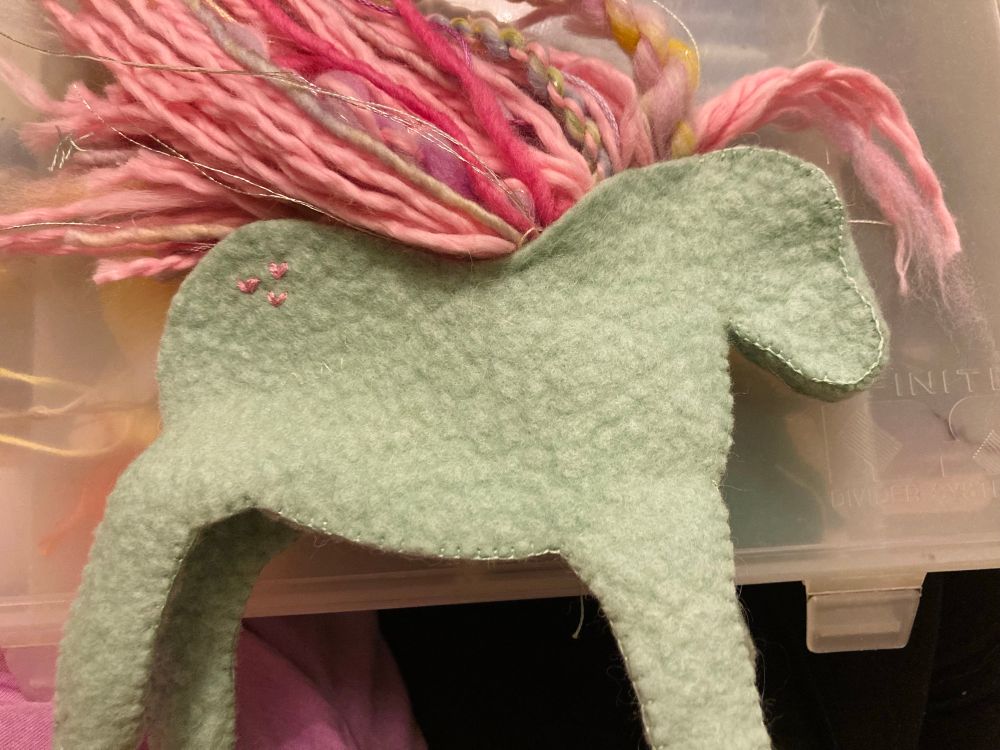 An in-progress picture of a unicorn I am sewing. It has a teal wool felt body with a mane made up of various textures and shades of pink yarn and wool roving. 