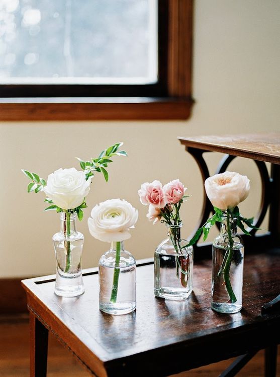 Four small glass vases each hold a single stem of flowers, including white roses and pink blooms, on a wooden table. A window with a blurred view is in the background.