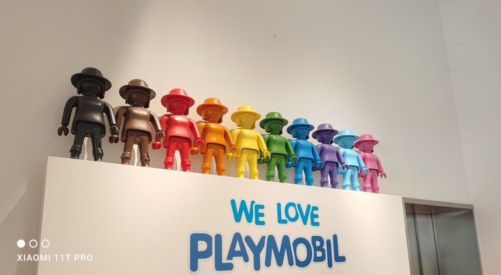 Eine weiße Mauer auf der einfarbige, übergroße Playmobilfiguren nebeneinander stehen. Sie sind farblich, in Anlehnung an die Pride-Flag, sortiert.
Darunter steht "We Love Playmobil" in großen Buchstaben.