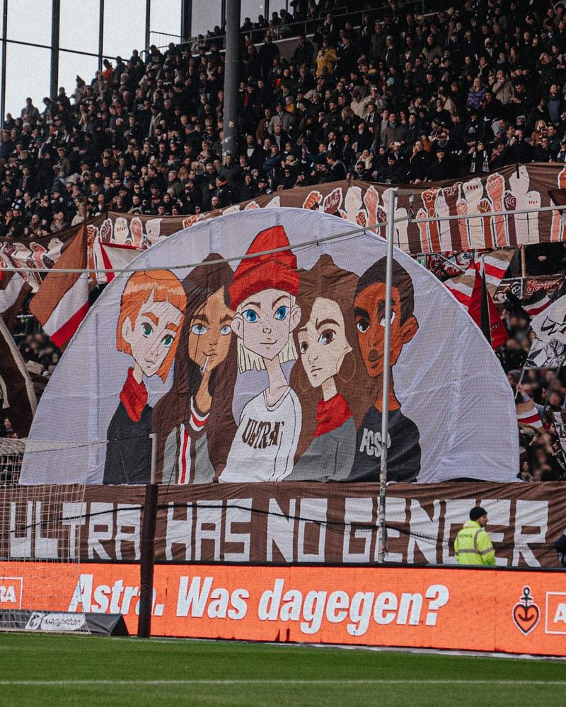The photo is of a large tifo at the Millerntor. In the foreground is a banner with the words "ULTRA HAS NO GENDER". Above it an artistic depiction of five different people in a modern cartoon style.