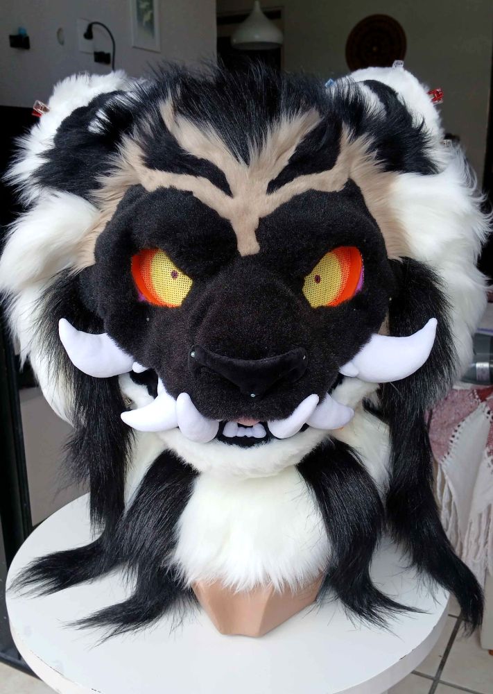 Fursuit head of a tiger with dark markings on most of its face, wide reddish eyes and teeth poking outwards similar to an oni mask. Some of its stripes extend out of the body and drapes onto the mannequin head it's mounted on.