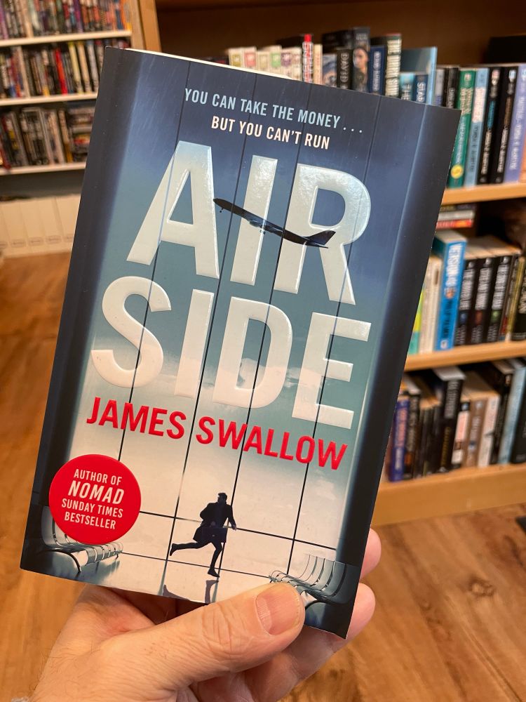 Trade paperback of Airside, cover featuring a man with a briefcase looking over his shoulder as he runs through an empty airport; through the large window behind him a plane has taken off and is high and climbing, wheels still down.