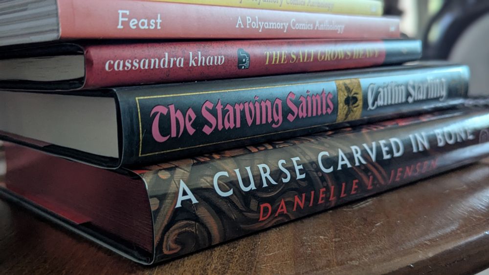 A stack of books: A Curse Carved in Bone by Danielle L. Jensen, The Starving Saints by Caitlin Starling, The Salt Grows Heavy by Cassandra Khaw, and Feast: A Polyamory comic anthology 