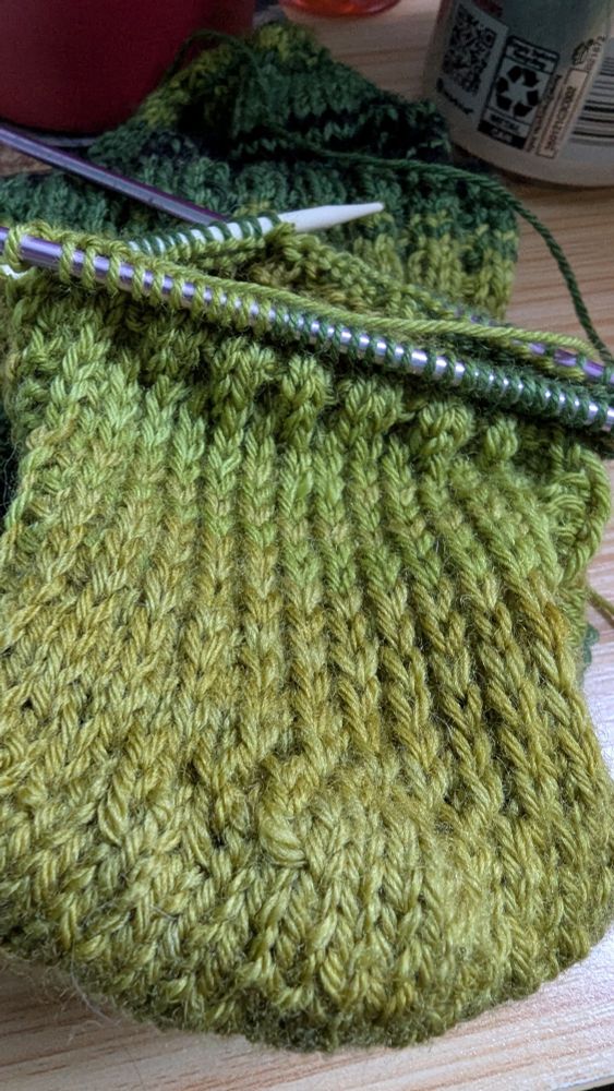 Heel flap of a sock being knit in a green and black striped yarn.