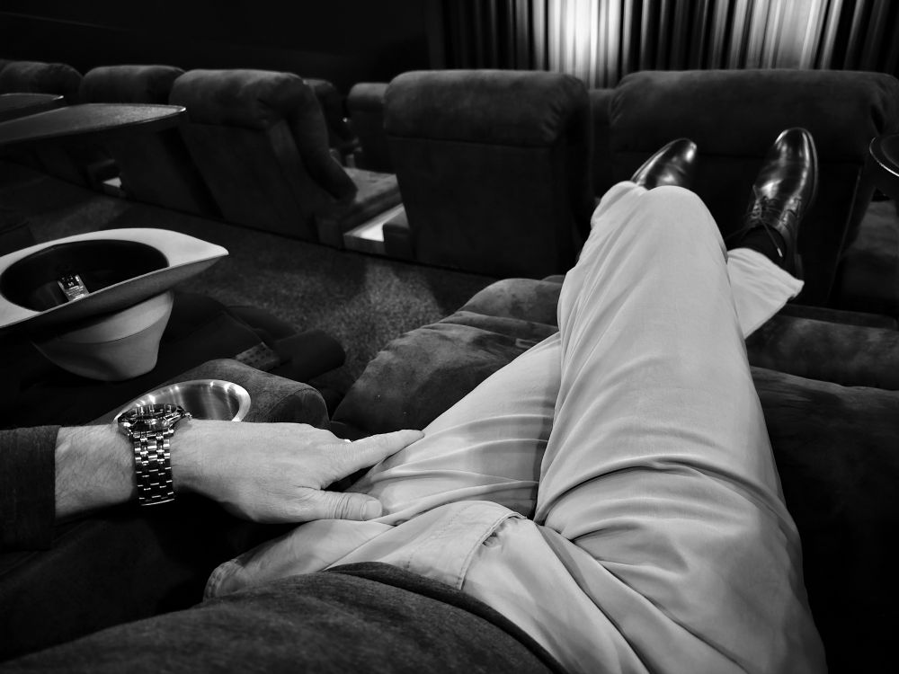 Ein Foto meiner unteren Körperhälfte, aufgenommen von leicht rechts. Ich sitze mit überschlagenen Beinen auf einem ausgefahrenen Liegesessel in einem Kinosaal. Links leben der Mittellehne liegt umgedreht ein grauer Fedora. Ich trage eine hellgraue Chino und ein dunkelgraues Oberteil. 