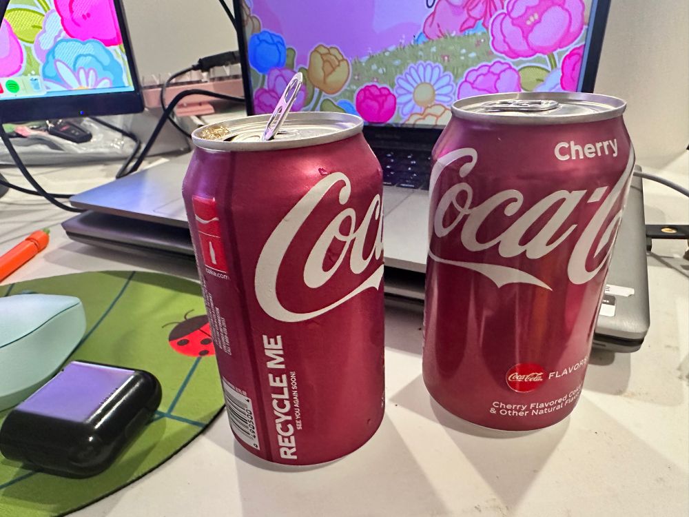 a photo of an opened can of cherry coke next to an unopened cherry coke. behind them is a laptop and some monitors, and to the left is a mouse on a mousepad next to an earbud holder.